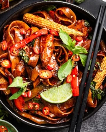 A bowl of stir-fried noodles with baby corn, red chilies, basil, red bell peppers, and a lime wedge, garnished with fresh herbs and served with black chopsticks.