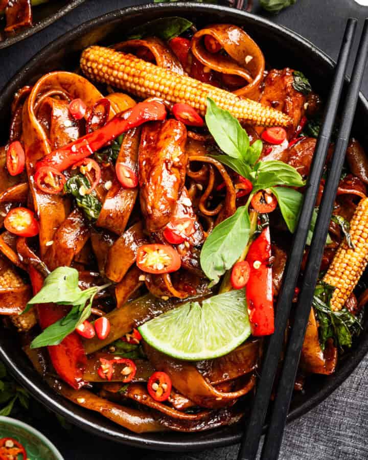 A bowl of stir-fried noodles with baby corn, red chilies, basil, red bell peppers, and a lime wedge, garnished with fresh herbs and served with black chopsticks.