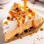 A slice of pumpkin pie with whipped cream, chopped pecans, and caramel sauce being drizzled on top, resting on a light-colored plate.