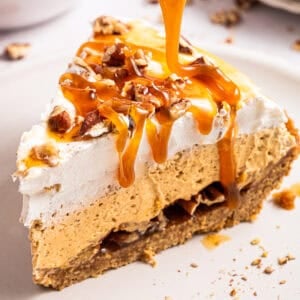 A slice of pumpkin pie with whipped cream, chopped pecans, and caramel sauce being drizzled on top, resting on a light-colored plate.