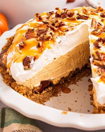 A pumpkin cream pie with whipped topping, caramel drizzle, and chopped pecans sits in a white pie dish. A large slice is missing, showing the creamy filling and graham cracker crust. Small pumpkins are in the background.