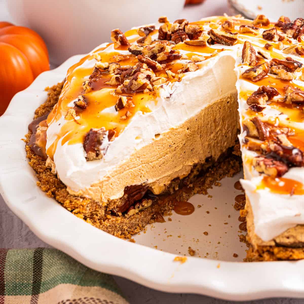 A pumpkin cream pie with whipped topping, caramel drizzle, and chopped pecans sits in a white pie dish. A large slice is missing, showing the creamy filling and graham cracker crust. Small pumpkins are in the background.