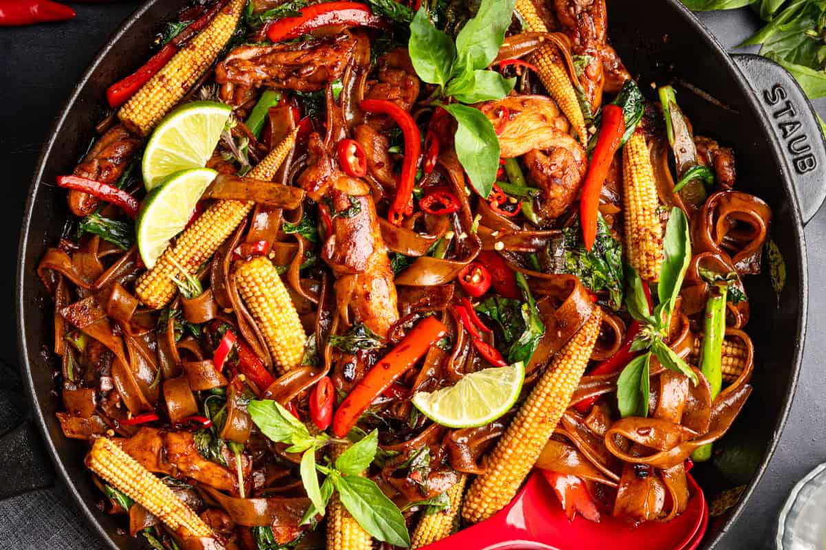 A skillet filled with stir-fried noodles, baby corn, red bell peppers, chicken, fresh basil leaves, and lime wedges, garnished with red chili slices.