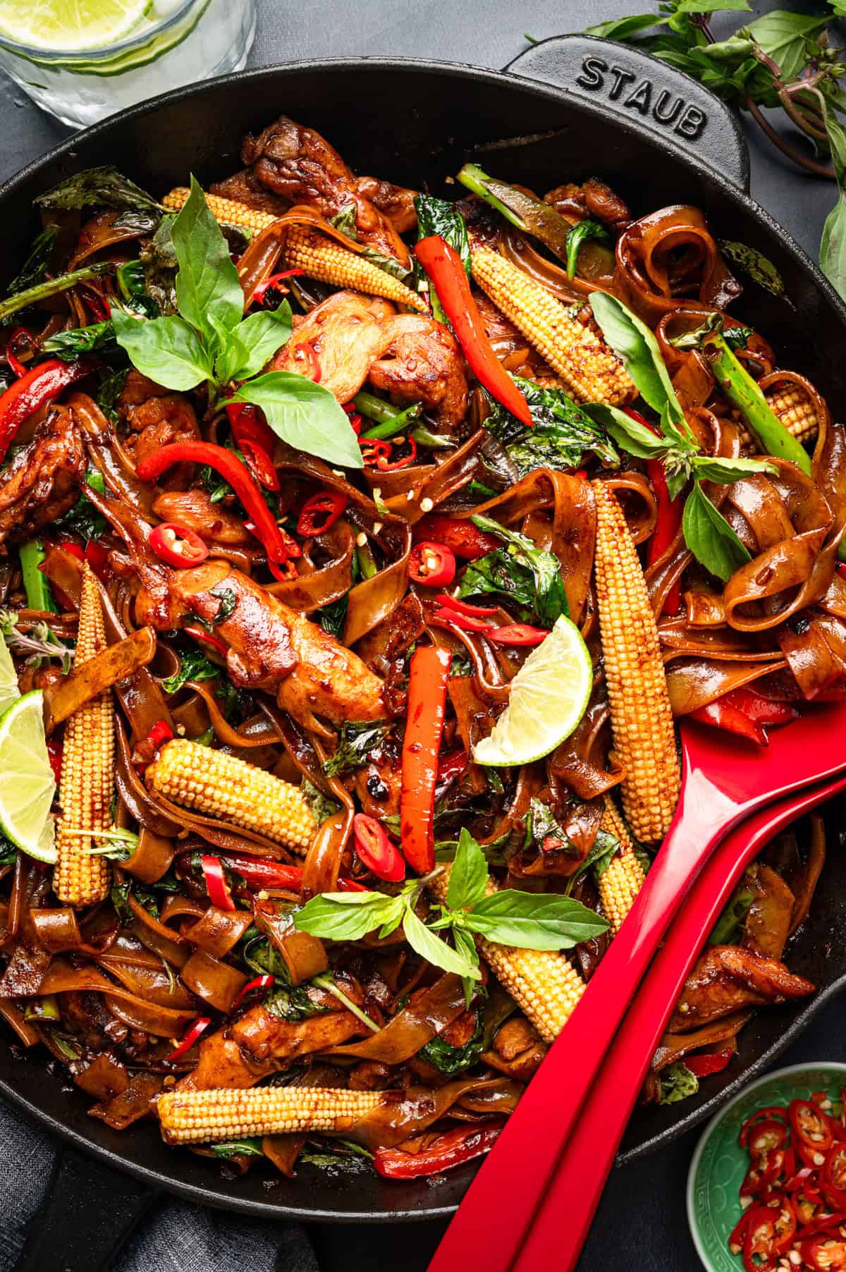 Thai Drunken Noodles A skillet filled with stir-fried noodles, baby corn, red peppers, chicken, basil, chili slices, and lime wedges, with a red serving spoon.