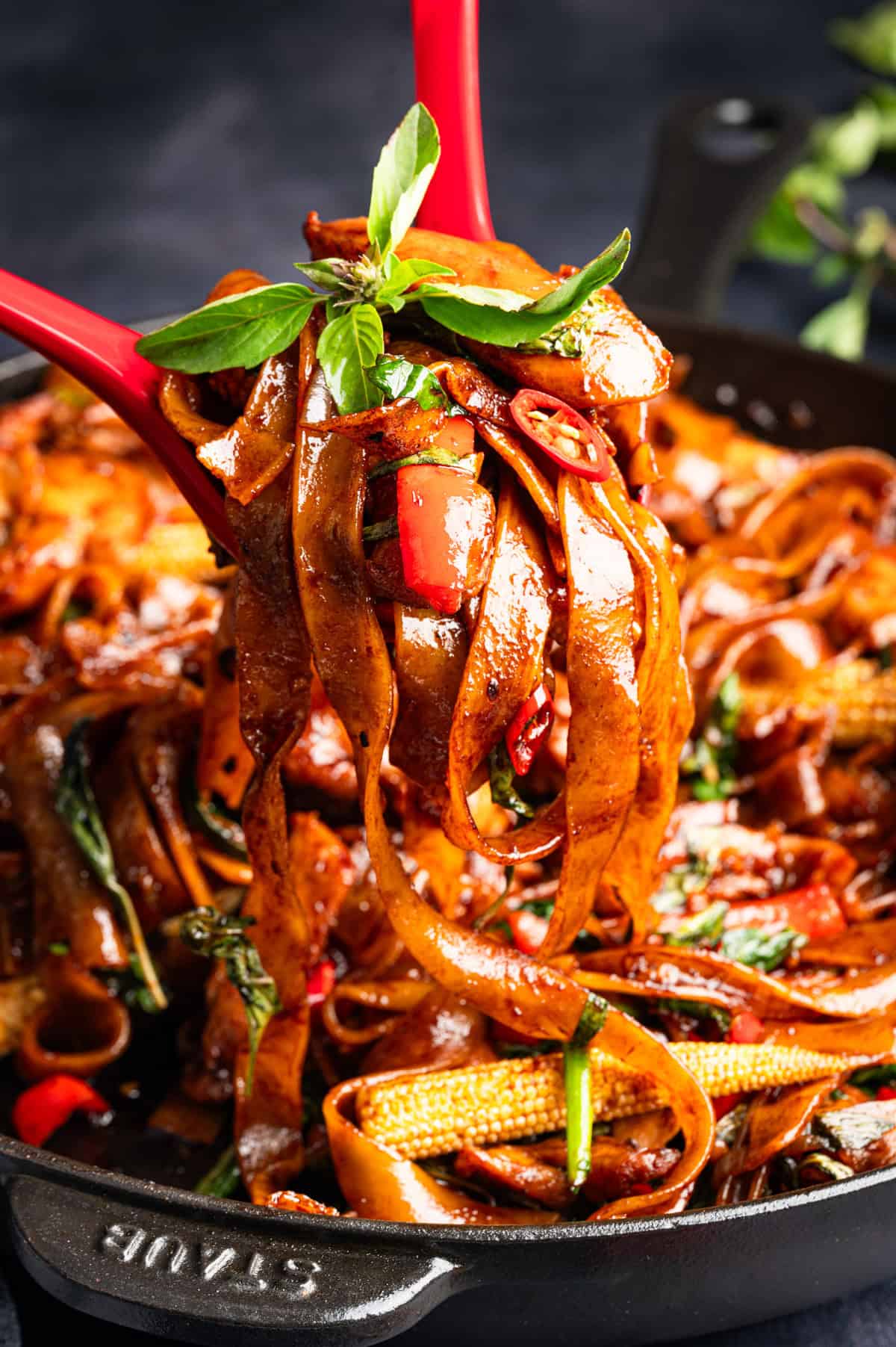 Thai Drunken Noodles Wide rice noodles stir-fried with vegetables, red chilies, and basil, lifted from a black skillet with red tongs.