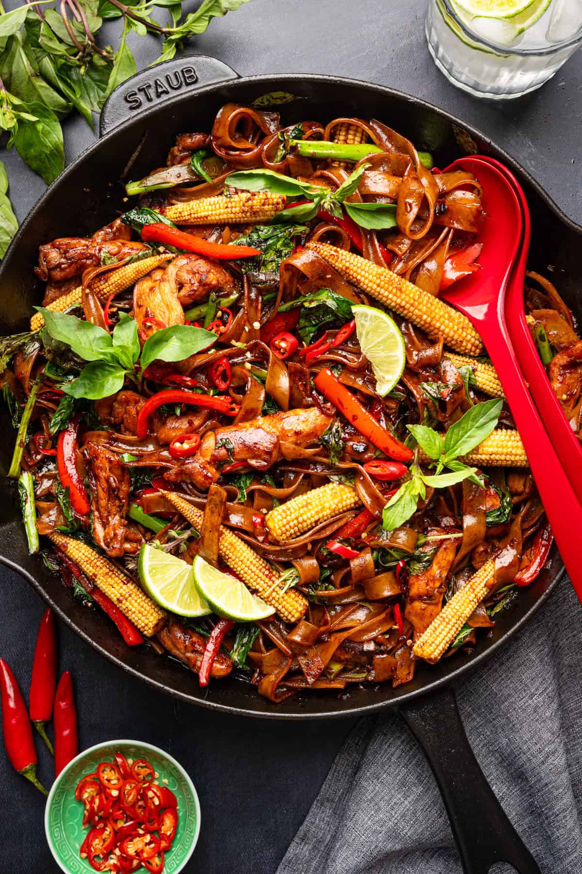 Thai Drunken Noodles A skillet filled with stir-fried noodles, chicken, baby corn, red chilies, basil, and lime wedges, with a red serving spoon on the side.