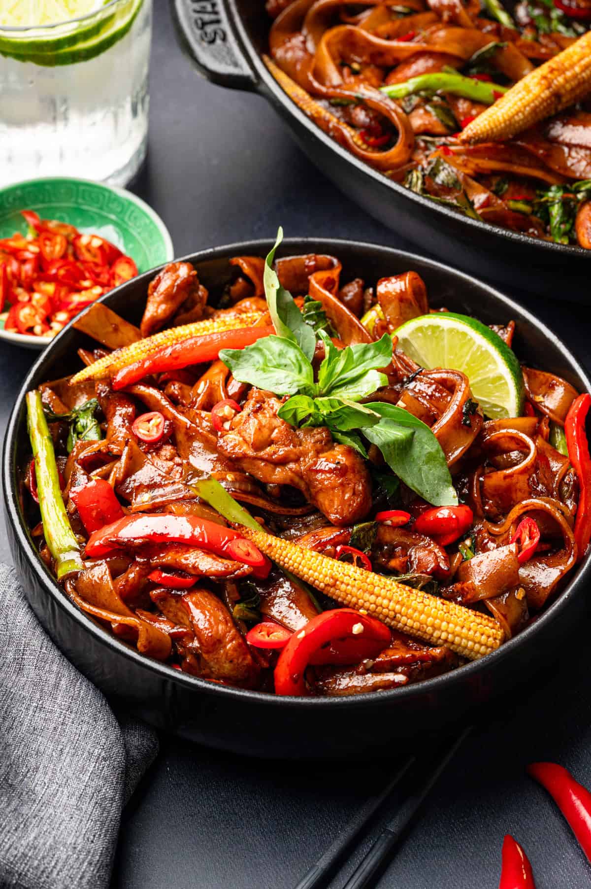 Thai Drunken Noodles A black bowl filled with stir-fried noodles, chicken, baby corn, red chilies, and green vegetables, garnished with fresh basil and a lime wedge.