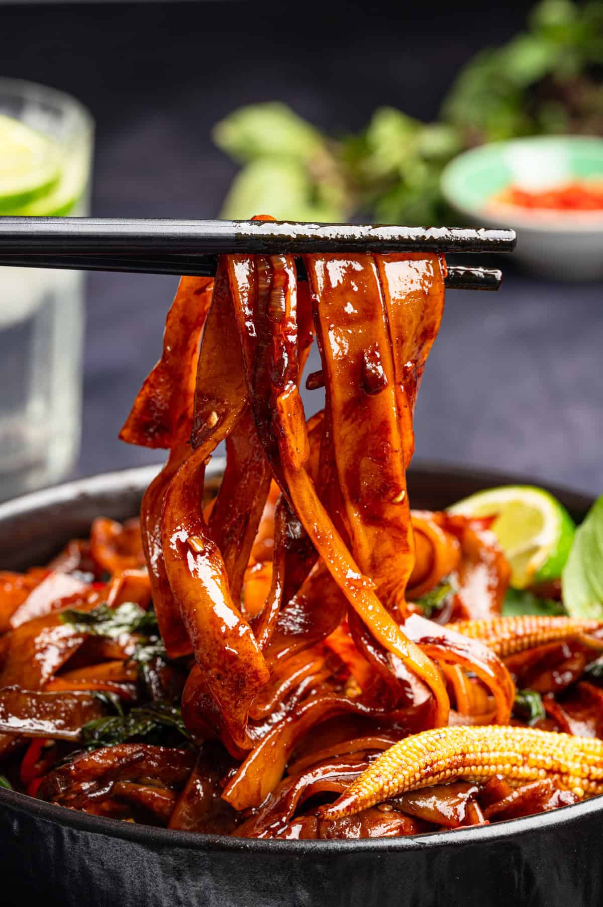 Thai Drunken Noodles Chopsticks lift wide, glossy noodles coated in a dark sauce from a bowl with vegetables like baby corn and greens.