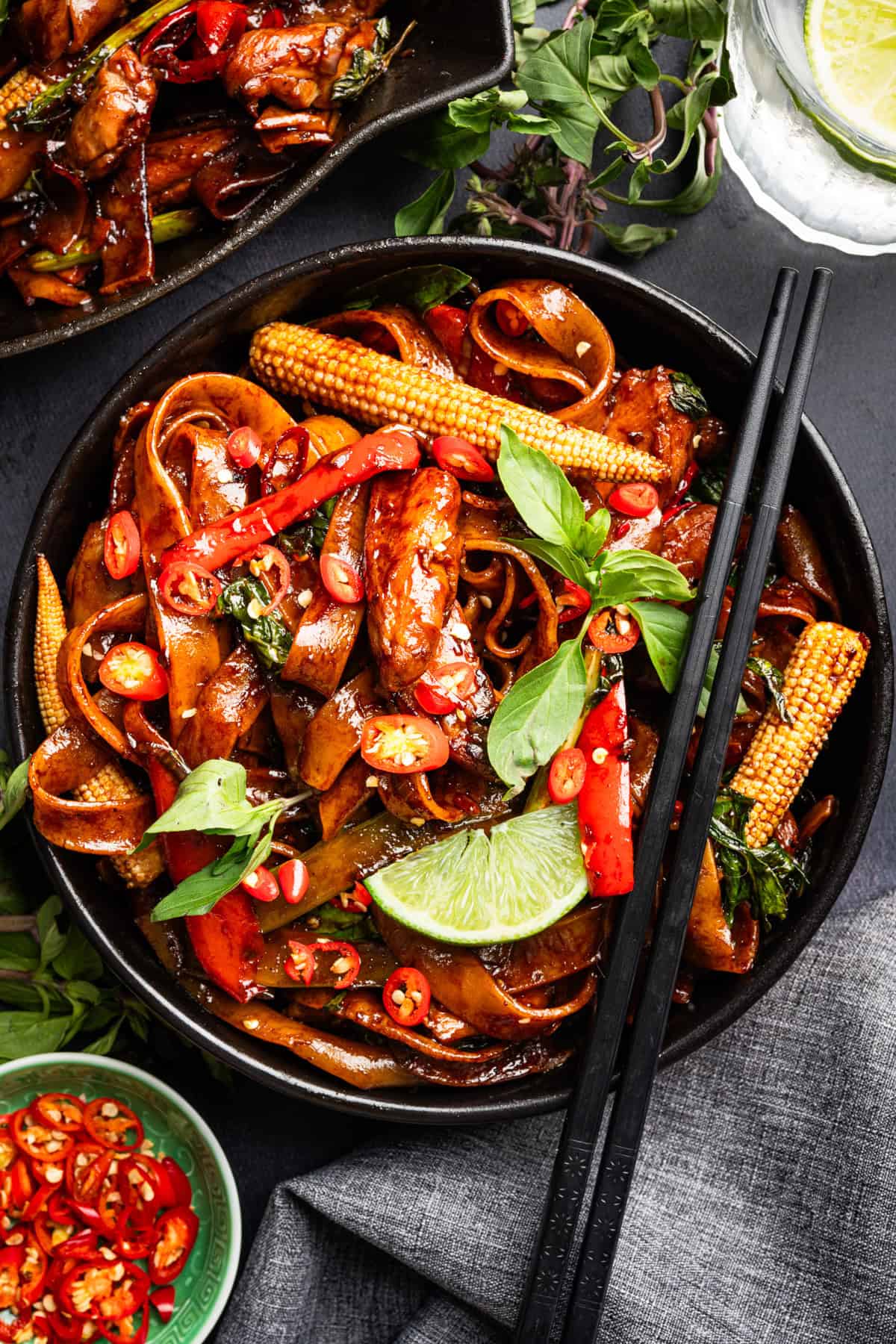Thai Drunken Noodles A black bowl of stir-fried noodles with chicken, baby corn, red chili, basil, and lime wedge.