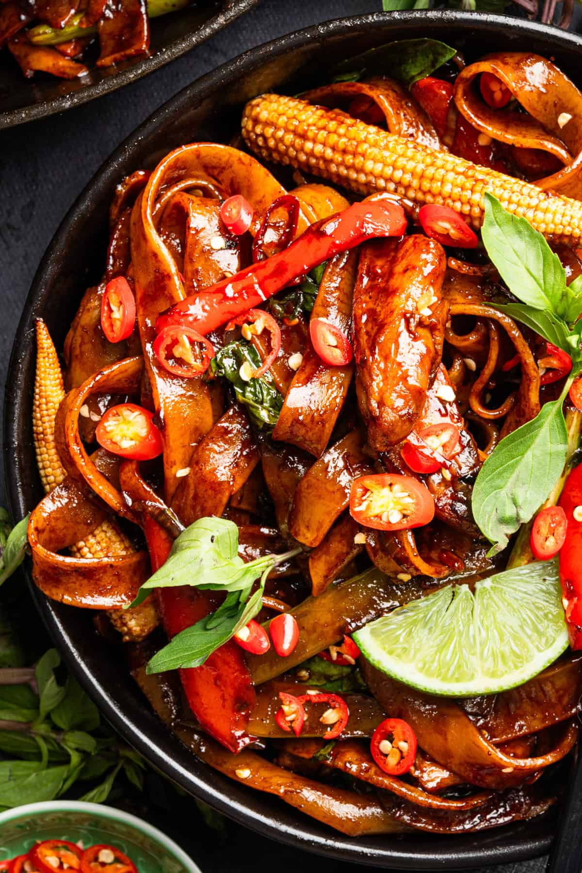 Thai Drunken Noodles A bowl of spicy stir-fried noodles with sliced red chili, baby corn, basil leaves, and a lime wedge, garnished with sauce and vegetables.