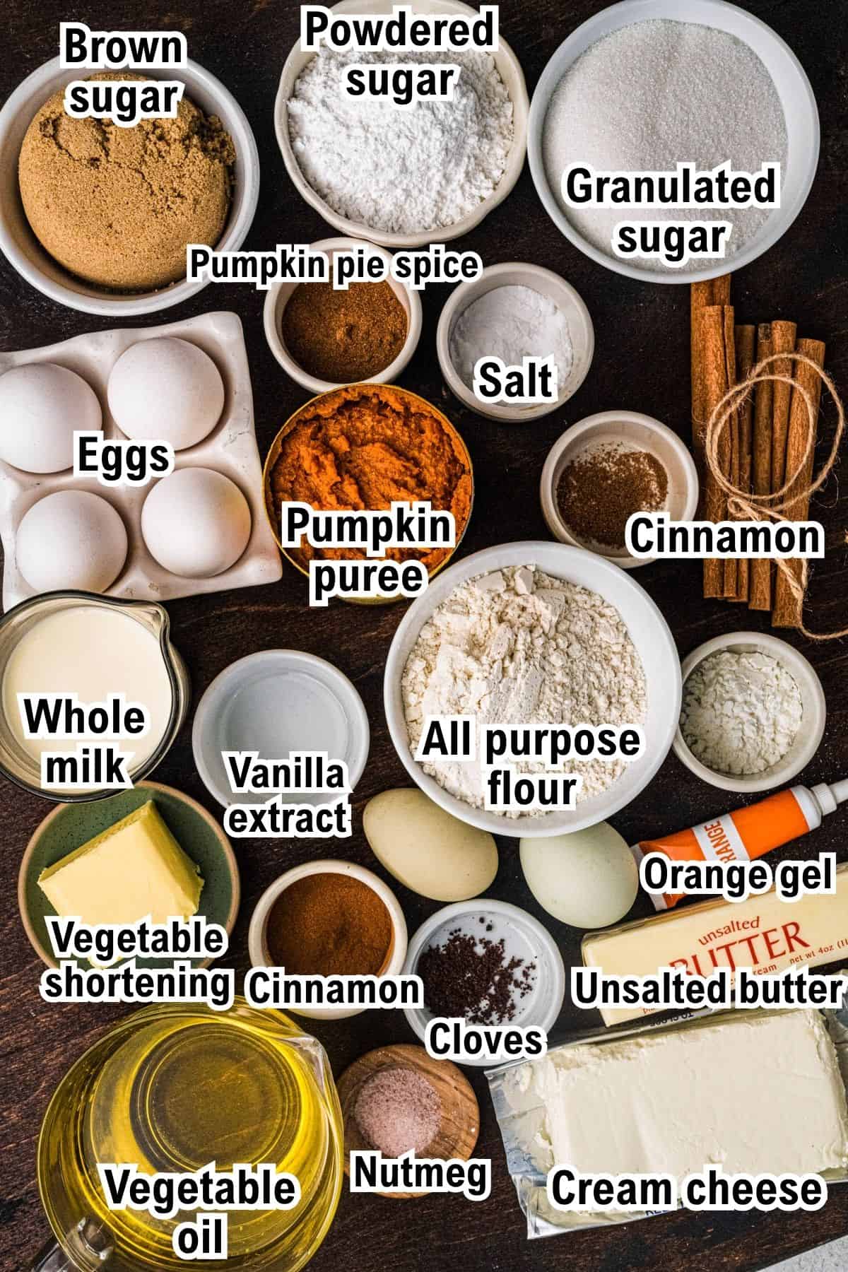 The Ingredients for Pumpkin Shaped Cake This photo shows the ingredients for Pumpkin Shaped Cake including sugars, eggs, pumpkin puree, spices, flour, whole milk, vegetable oil, shortening, butter, orange gel, and cream cheese on a dark surface.