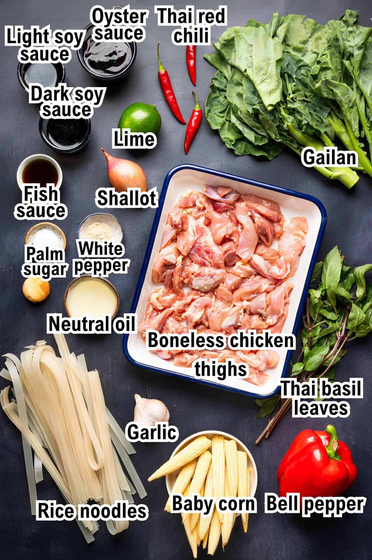 The Ingredients for Thai Drunken Noodles This photo shows the Ingredients for Thai Drunken Noodles: labeled sauces, herbs, vegetables, rice noodles, and raw boneless chicken thighs in a tray, all on a dark surface.