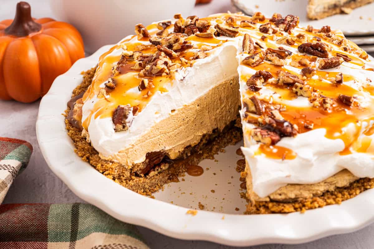 A creamy pie with a graham crust, topped with whipped cream, chopped pecans, and caramel drizzle. A slice is missing, revealing a light brown filling. 