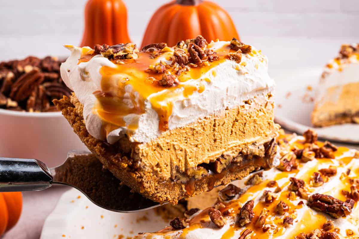 Turtle Pumpkin Pie A slice of pumpkin pie with whipped cream, caramel sauce, and pecans is being lifted from a pie.