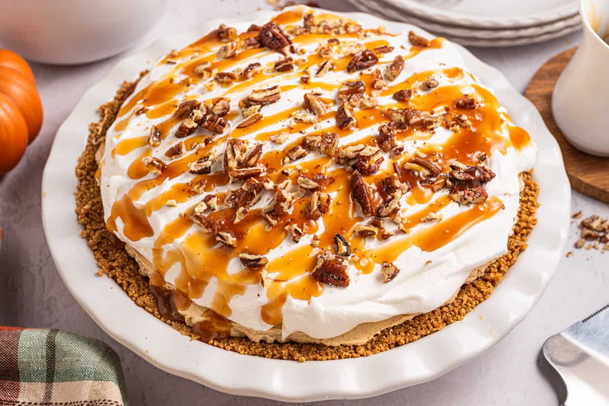 Turtle Pumpkin Pie A pie with a crumb crust, topped with whipped cream, chopped pecans, and caramel drizzle sits on a white pie dish.