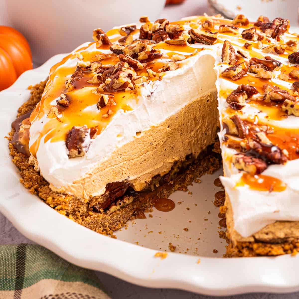 Turtle Pumpkin Pie A creamy pumpkin pie topped with whipped cream, caramel drizzle, and chopped pecans sits in a white dish with a slice removed, showing its layers.