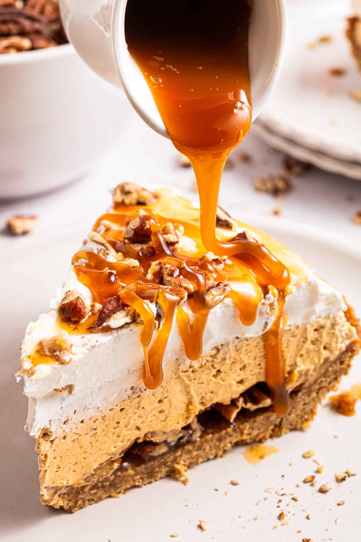 Turtle Pumpkin Pie A slice of pumpkin pie topped with whipped cream and chopped pecans is being drizzled with caramel sauce from a small pitcher.