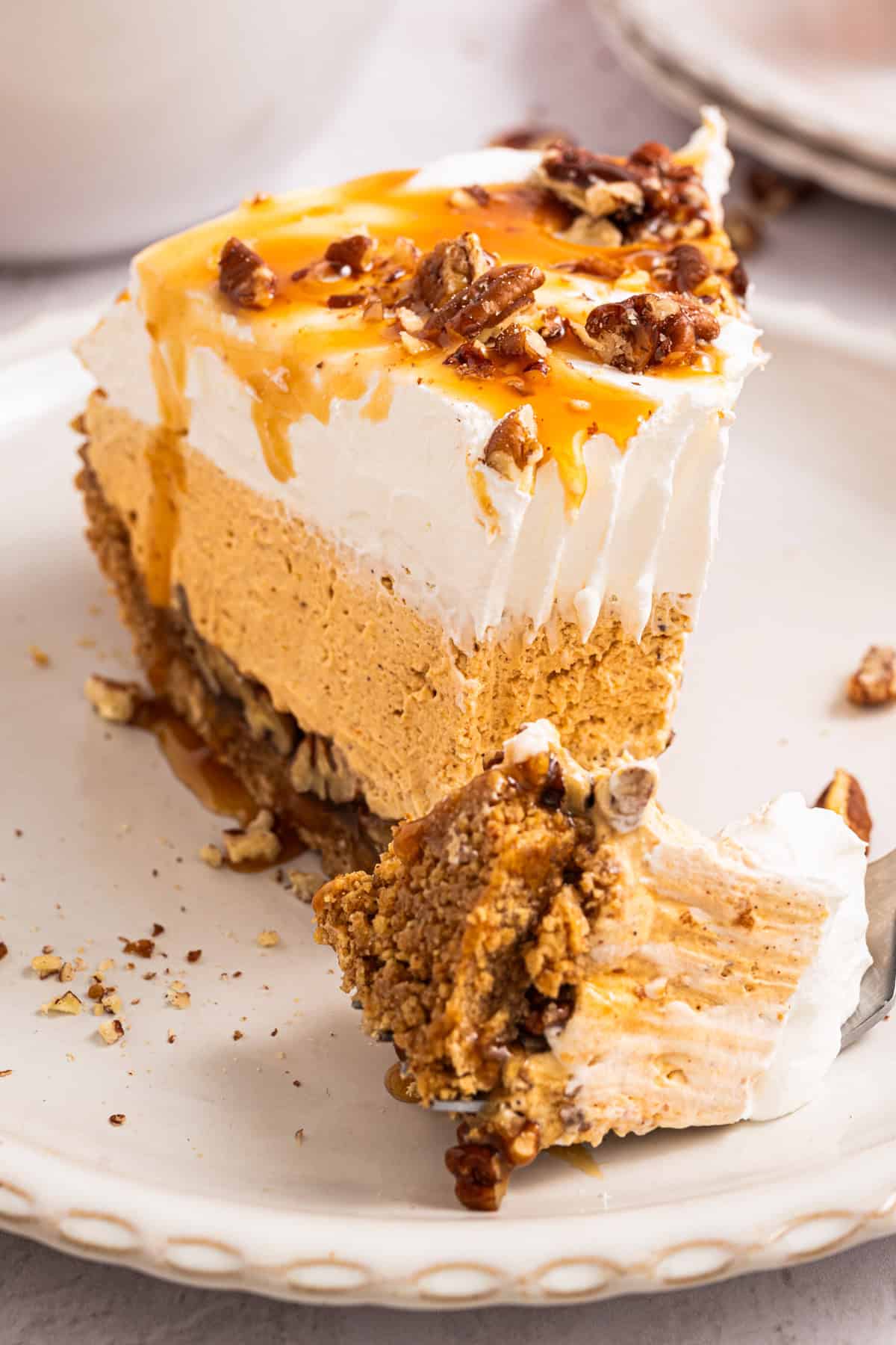 Turtle Pumpkin Pie A slice of layered pumpkin pie topped with whipped cream, caramel sauce, and chopped pecans, with a bite taken out on a fork, sits on a white plate.