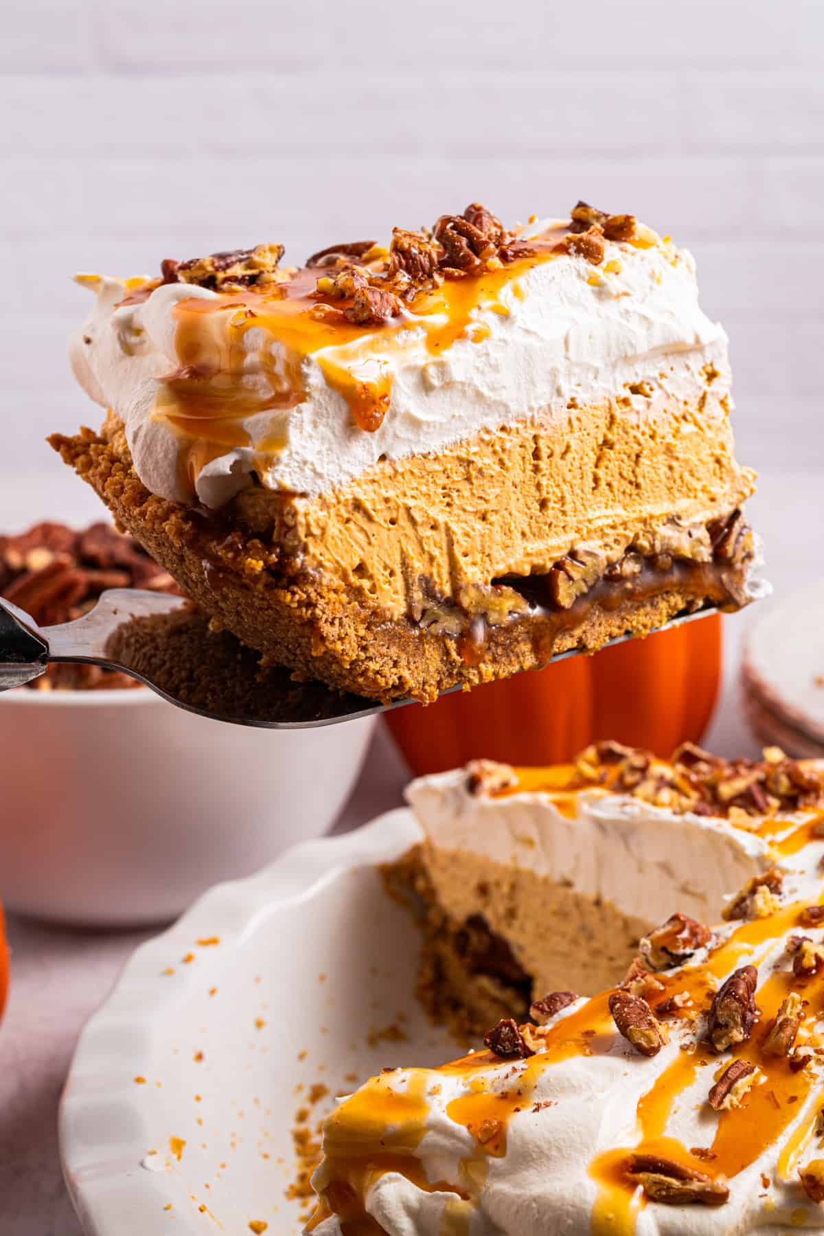 Turtle Pumpkin Pie A slice of creamy pumpkin pie topped with whipped cream, caramel sauce, and pecans is being lifted from a whole pie.