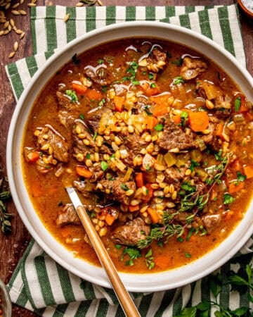 A bowl of beef and barley soup with carrots and fresh herbs, served with a spoon on a striped cloth napkin.