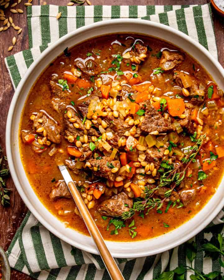 A bowl of beef and barley soup with carrots and fresh herbs, served with a spoon on a striped cloth napkin.