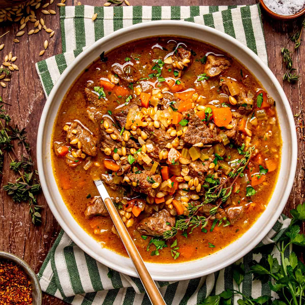 A bowl of beef and barley soup with carrots and fresh herbs, served with a spoon on a striped cloth napkin.