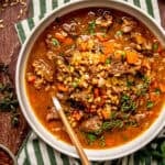 A bowl of beef and barley soup with carrots and fresh herbs, served with a spoon.