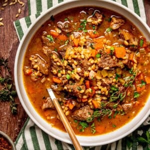 A bowl of beef and barley soup with carrots and fresh herbs, served with a spoon.