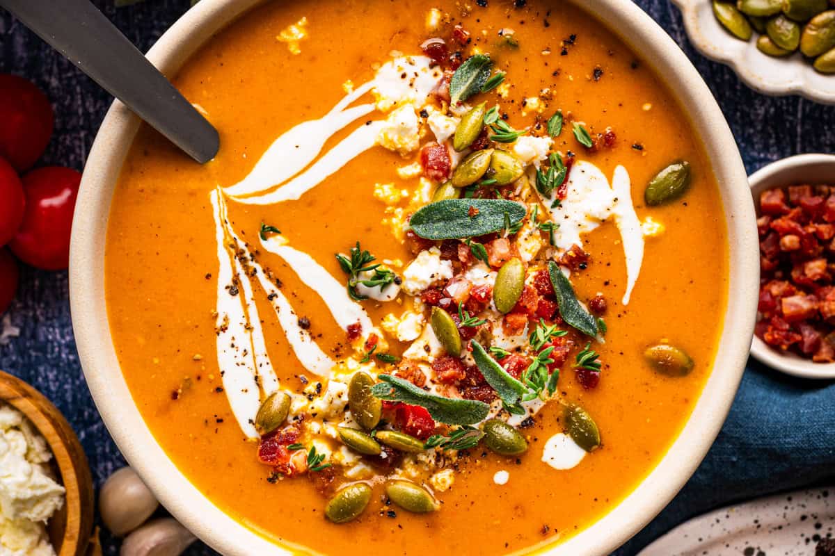 Butternut Squash Soup A bowl of orange soup garnished with cream swirls, crumbled cheese, bacon bits, pumpkin seeds, and fresh herbs.