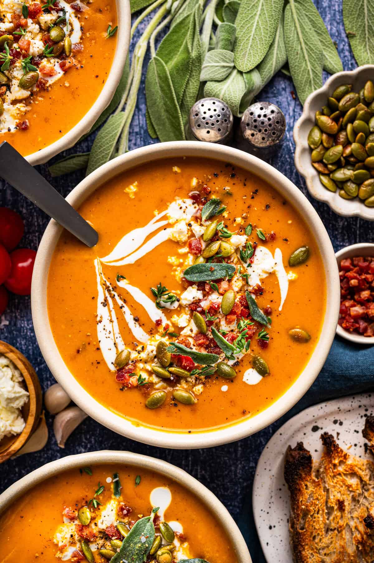 Butternut Squash Soup Bowls of creamy orange soup topped with swirls of cream, crumbled cheese, bacon bits, pumpkin seeds, and fresh sage leaves, served with toasted bread.