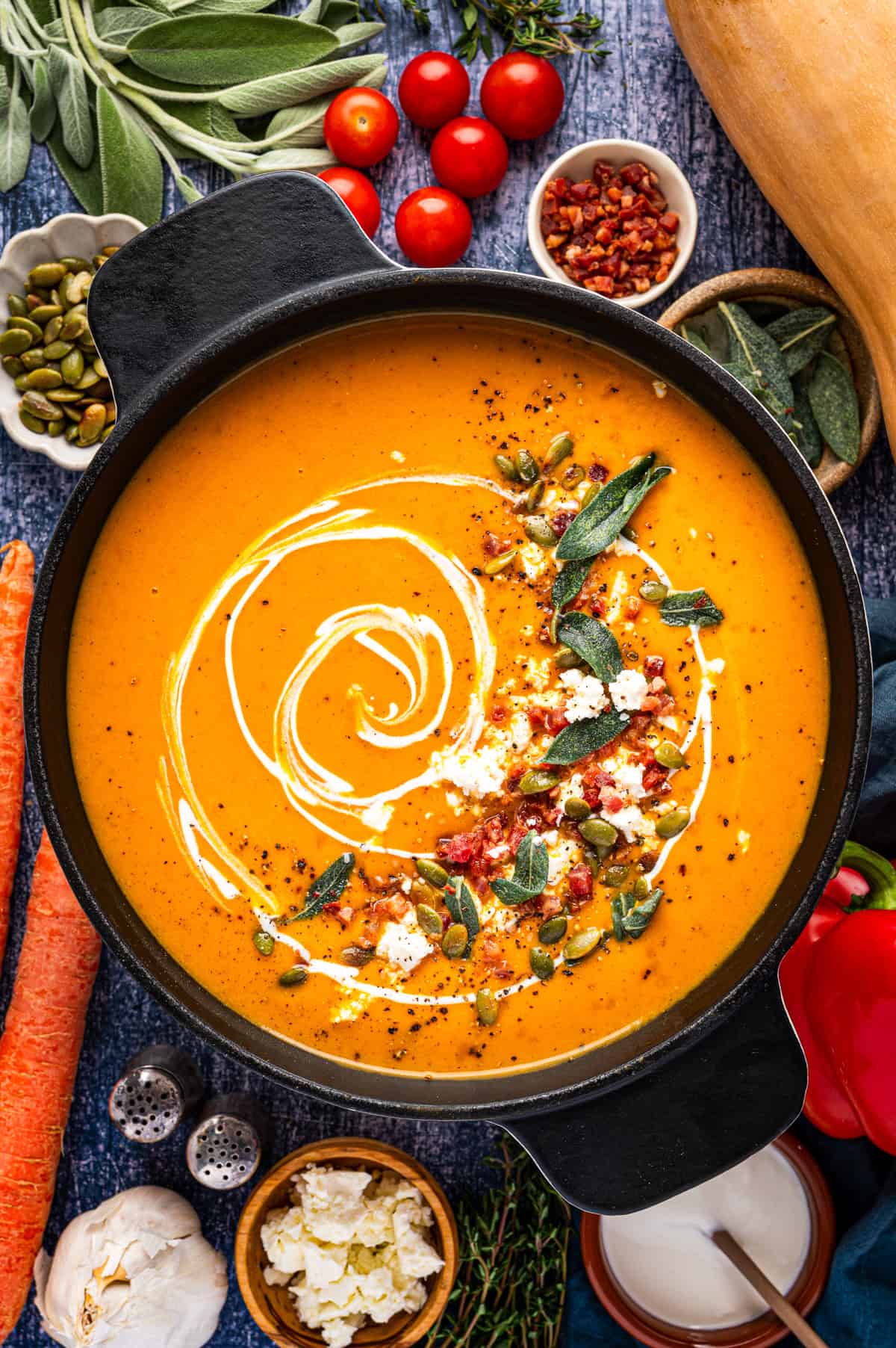 Butternut Squash Soup A black pot of orange soup swirled with cream and topped with pumpkin seeds, meat, cheese, and sage.