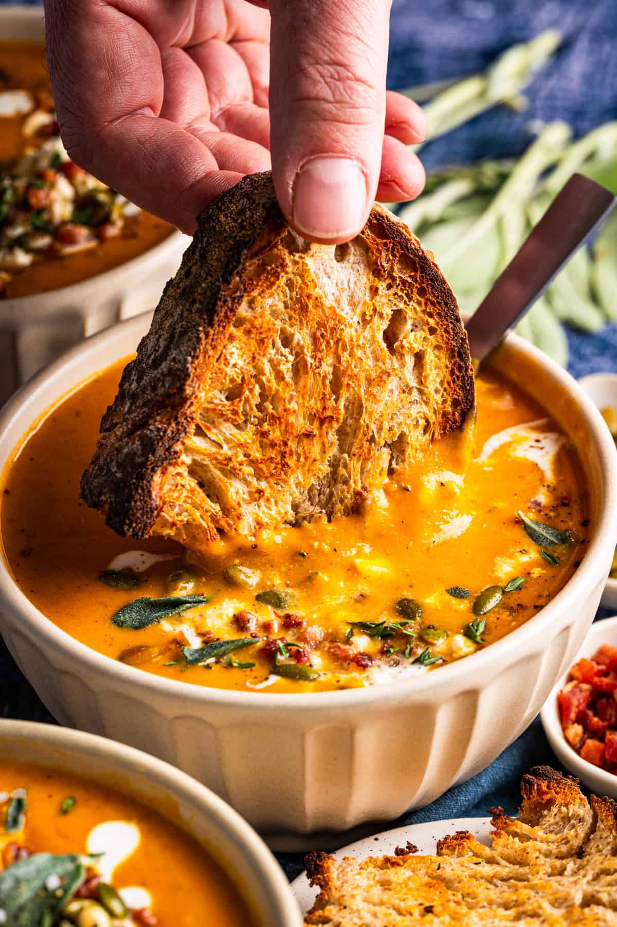 Butternut Squash Soup A hand dips a piece of toasted crusty bread into a bowl of creamy orange soup topped with herbs and bacon bits.