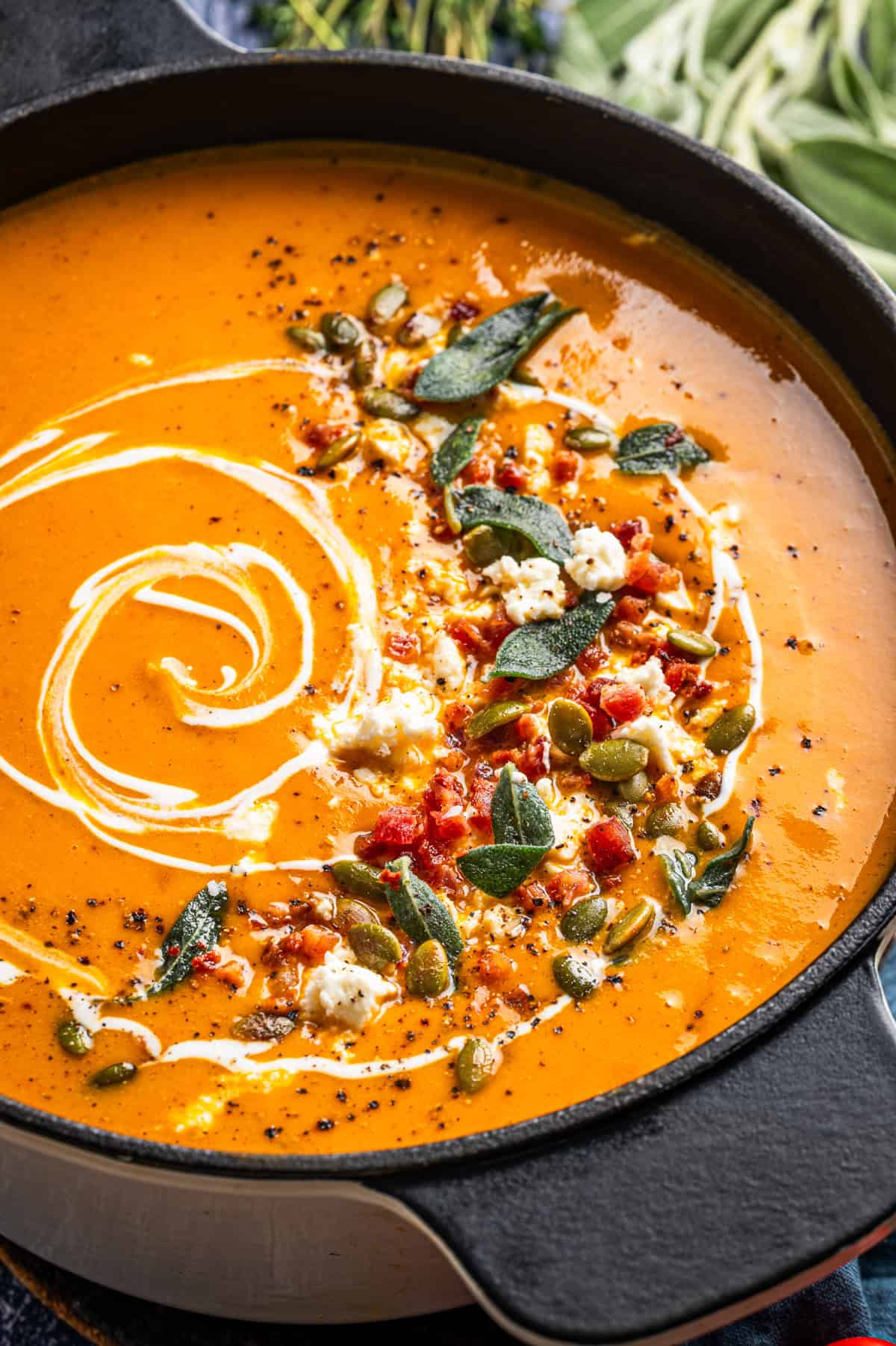 Butternut Squash Soup Creamy orange soup in a cast-iron pan, garnished with a swirl of cream, pumpkin seeds, crumbled cheese, bacon bits, and sage leaves.