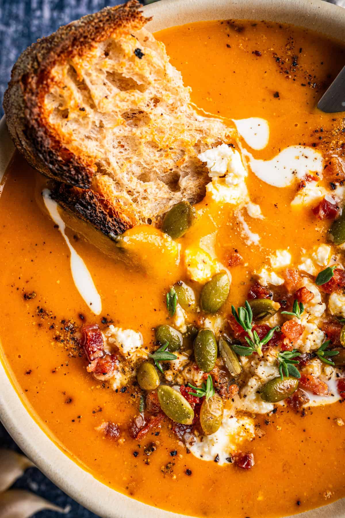Butternut Squash Soup A bowl of orange soup topped with pumpkin seeds, crumbled cheese, bacon bits, and herbs, with a slice of toasted bread dipped inside.