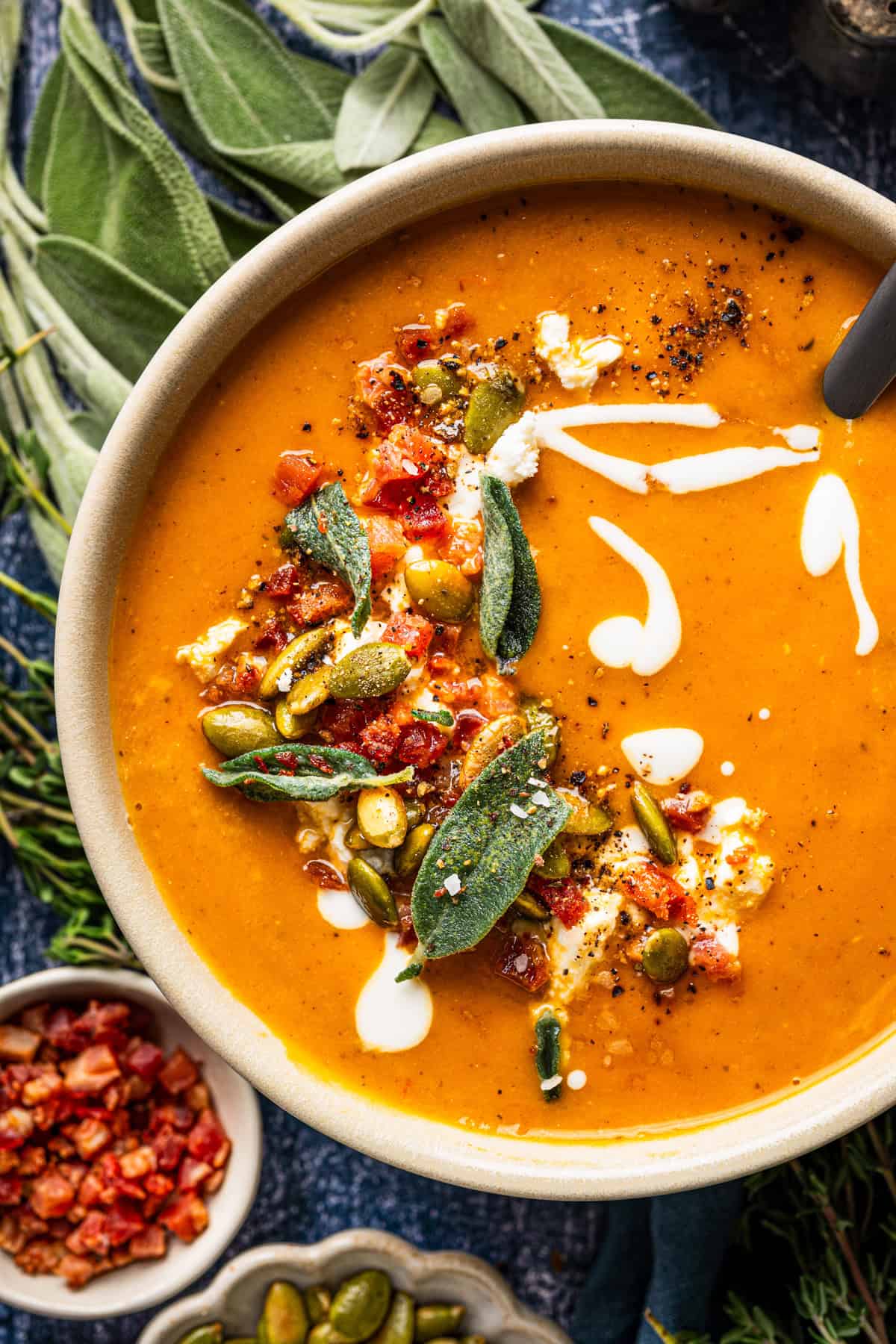 Butternut Squash Soup A bowl of orange soup topped with fried sage leaves, pumpkin seeds, bacon bits, and swirls of cream.