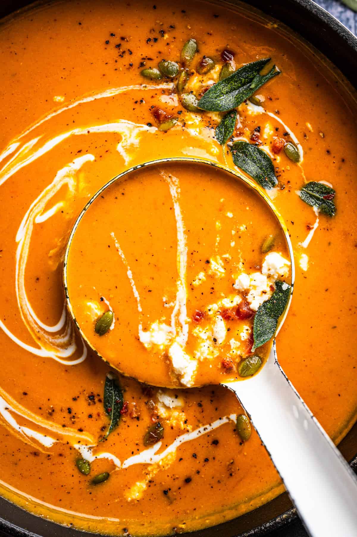 Butternut Squash Soup A ladle filled with creamy orange soup topped with pumpkin seeds, crumbled cheese, and fried sage leaves, resting in a larger pot of the same soup.
