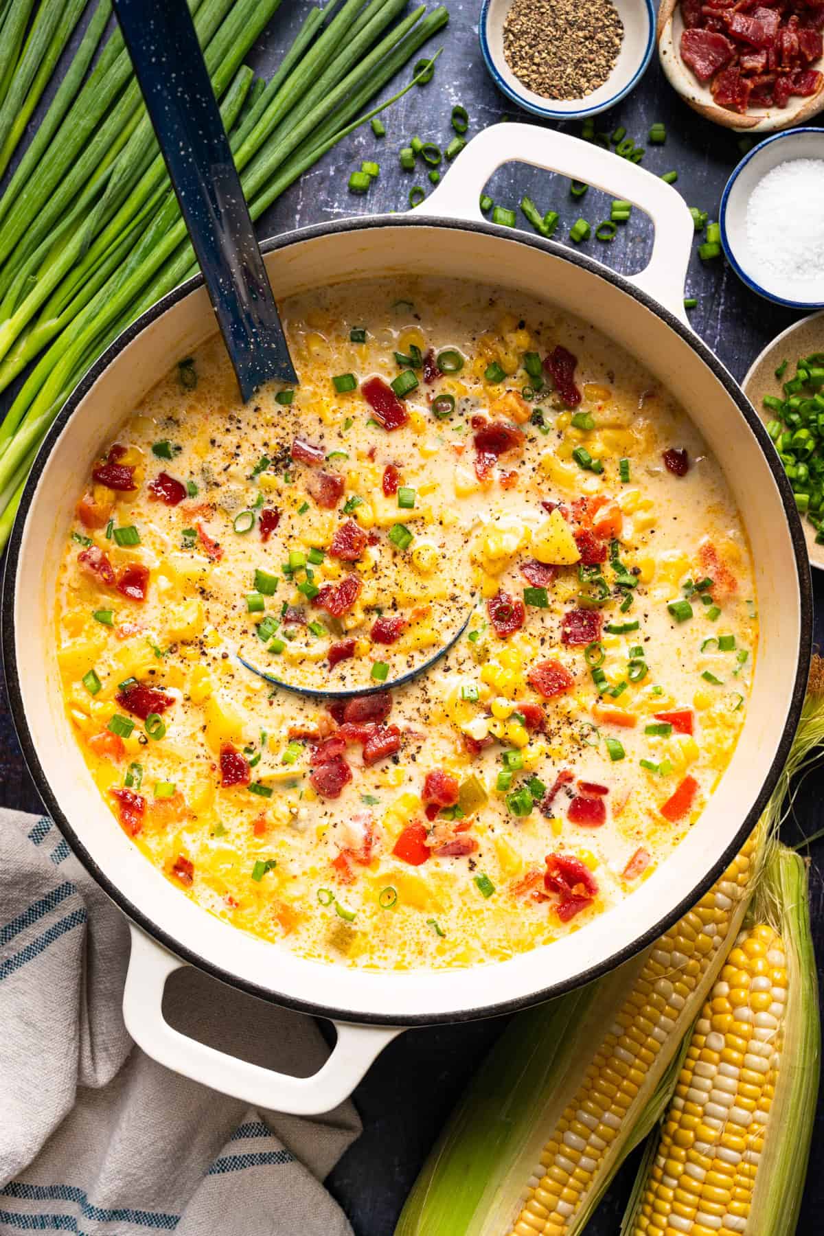 Corn Chowder with Bacon and Potatoes (One Pot) A large white pot filled with creamy corn chowder topped with bacon bits, green onions, and black pepper, with a ladle resting inside and fresh ears of corn nearby.