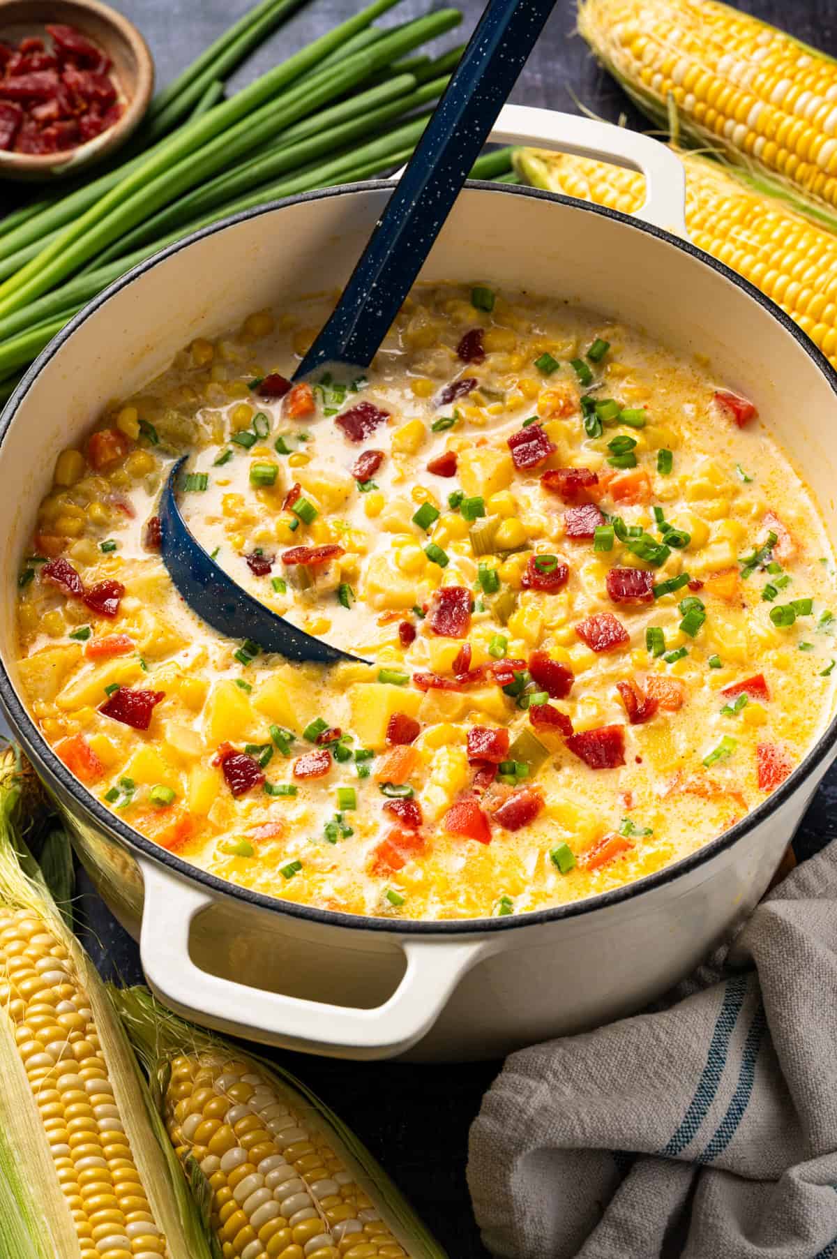 Corn Chowder with Bacon and Potatoes (One Pot) Creamy corn chowder in a white dutch oven, topped with bacon and chives, with a ladle inside and ears of corn nearby.