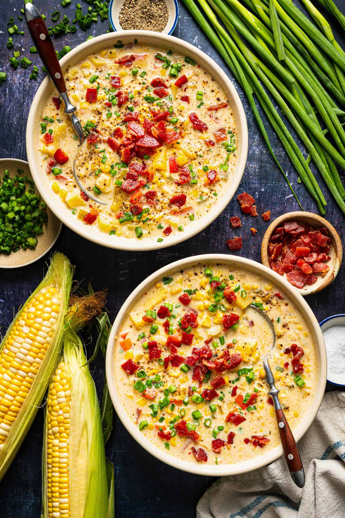 Corn Chowder with Bacon and Potatoes (One Pot) Two bowls of corn chowder topped with crumbled bacon and green onions, accompanied by ears of corn, a small bowl of bacon bits, and bowls of chopped onions, salt, and pepper.