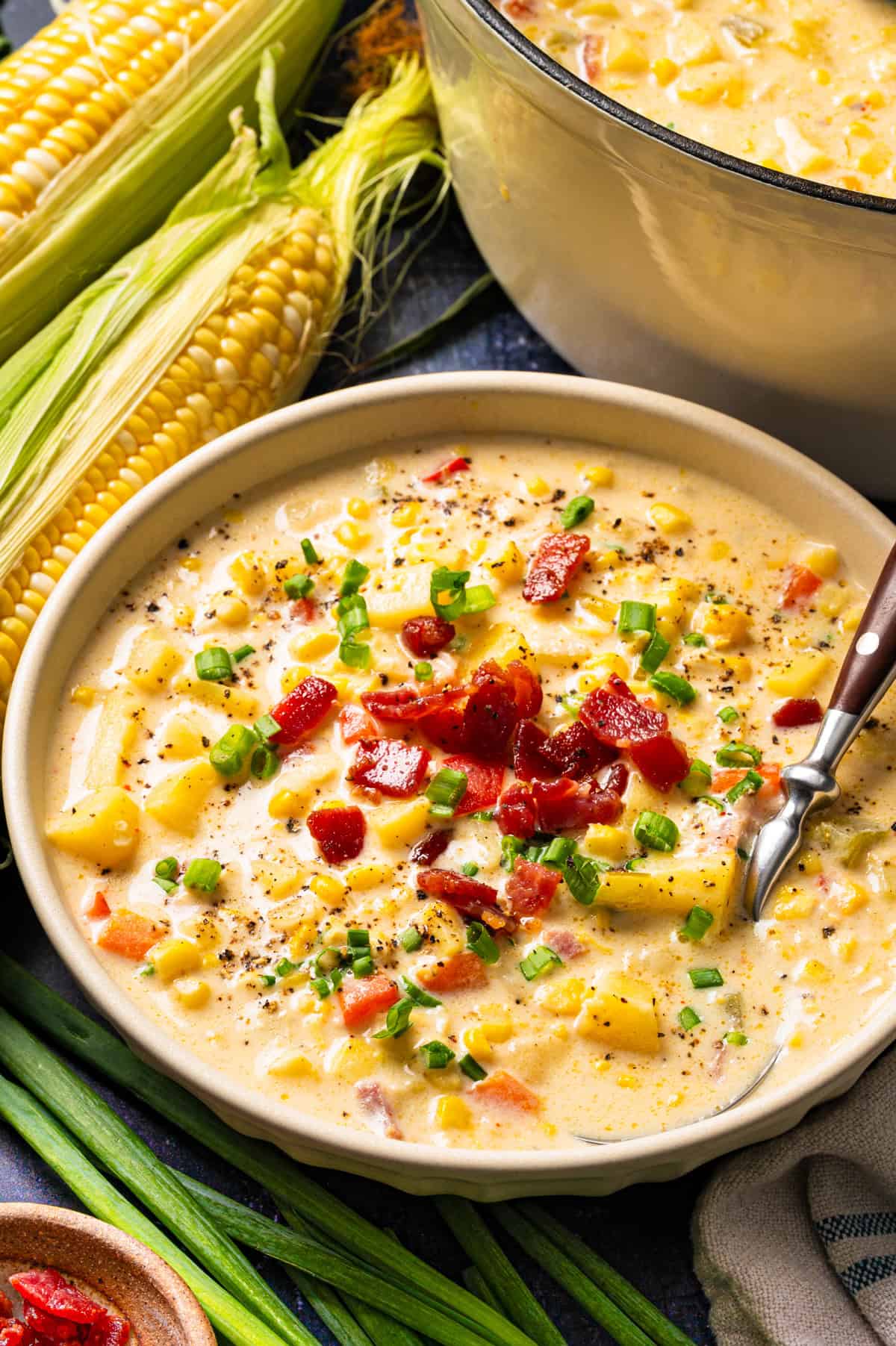 Corn Chowder with Bacon and Potatoes (One Pot) A bowl of creamy corn chowder topped with bacon and green onions, with ears of fresh corn and a large pot of soup in the background.