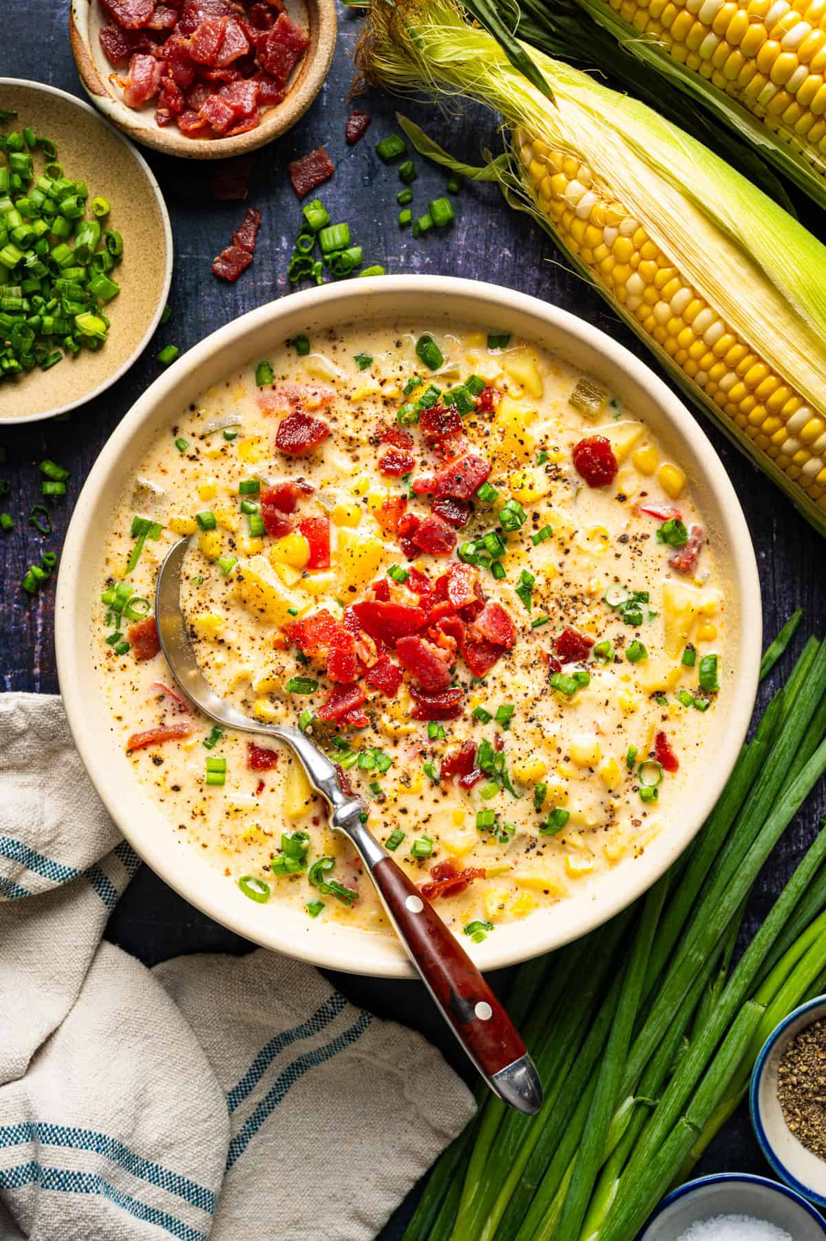 Corn Chowder with Bacon and Potatoes (One Pot) A bowl of creamy corn chowder topped with bacon and green onions, served with a spoon and fresh corn on the cob.