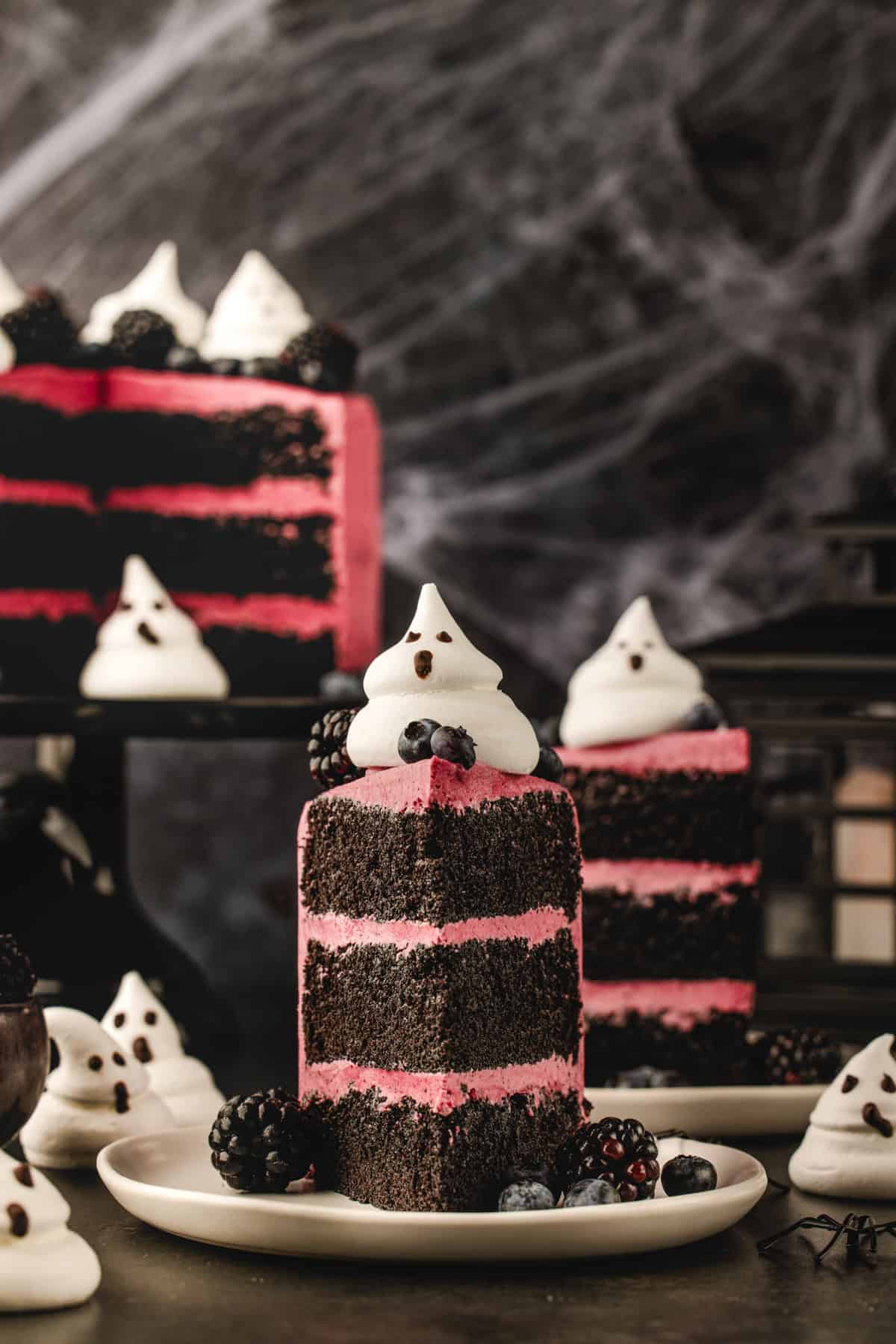 Halloween Cake A slice of chocolate cake with pink frosting, topped with a ghost-shaped meringue, on a plate with blackberries and blueberries.