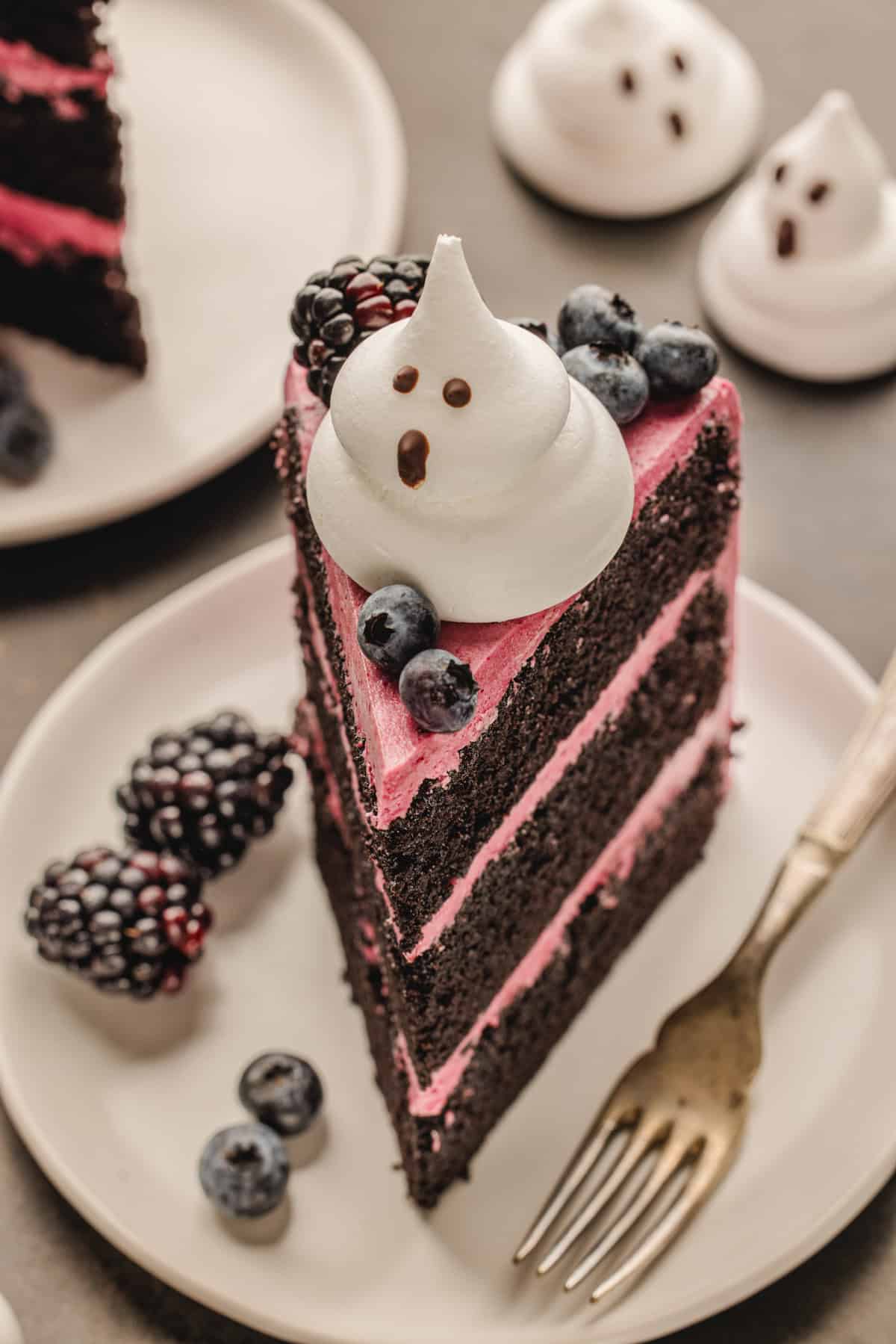 Halloween Cake A slice of chocolate layer cake with pink frosting, topped with a ghost-shaped meringue, blackberries, and blueberries.