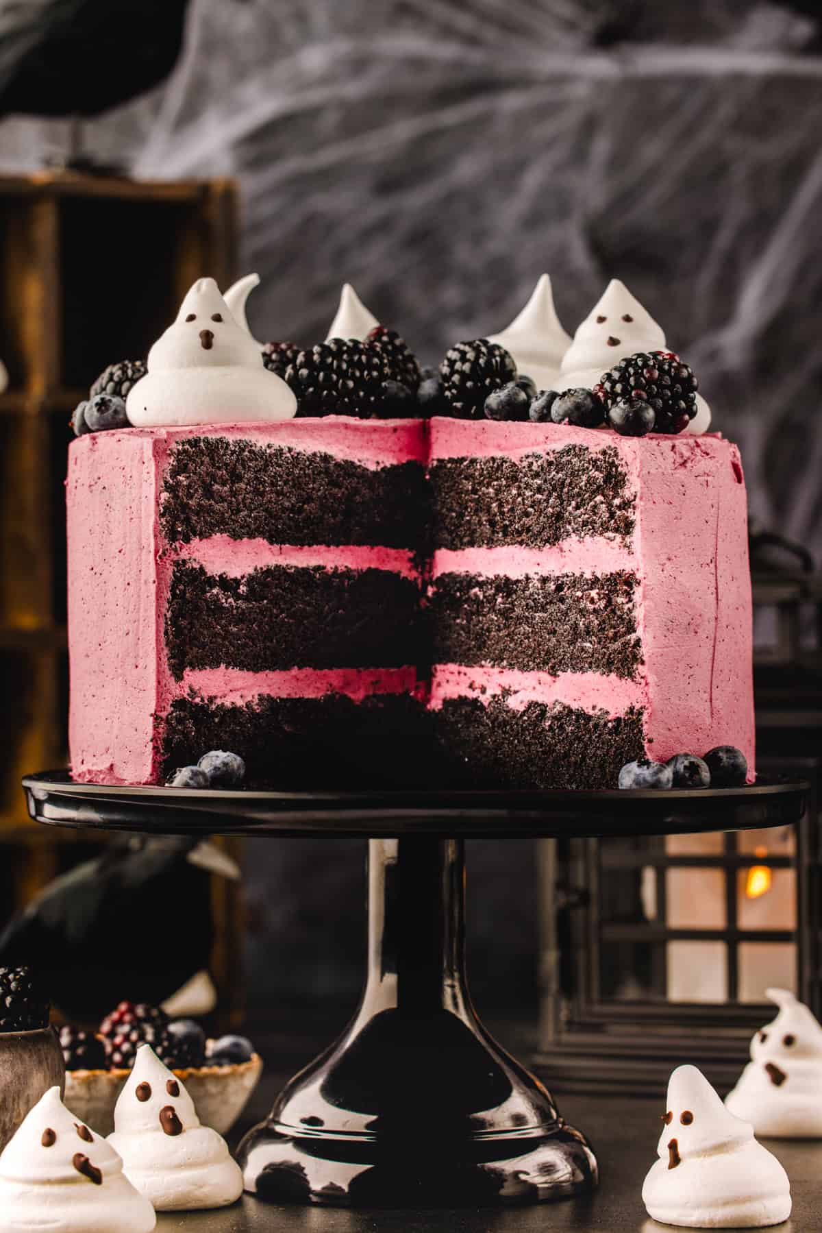 Halloween Cake A three-layer chocolate cake with pink frosting, topped with blackberries, blueberries, and ghost-shaped meringues, with a slice removed.