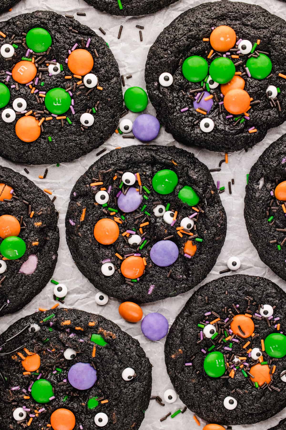 Halloween Cookies Several round, dark chocolate cookies decorated with colorful candy-coated chocolates, candy eyes, and sprinkles on parchment paper.