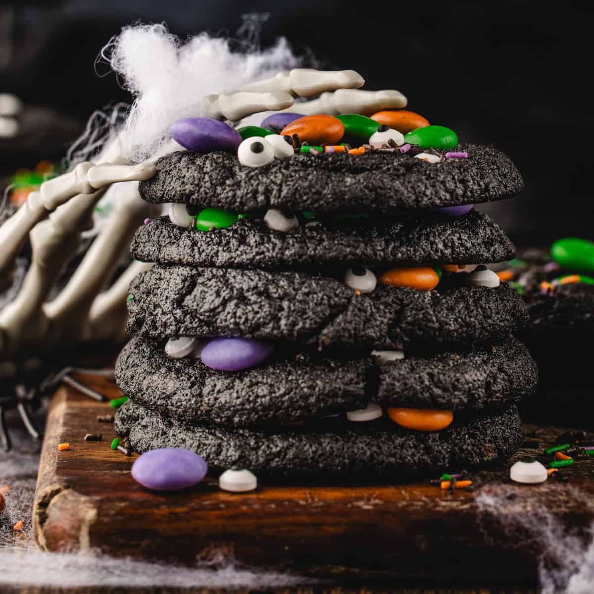 Halloween Cookies A stack of black Halloween cookies studded with candy eyes and colorful chocolate candies, with a skeletal hand resting on top.