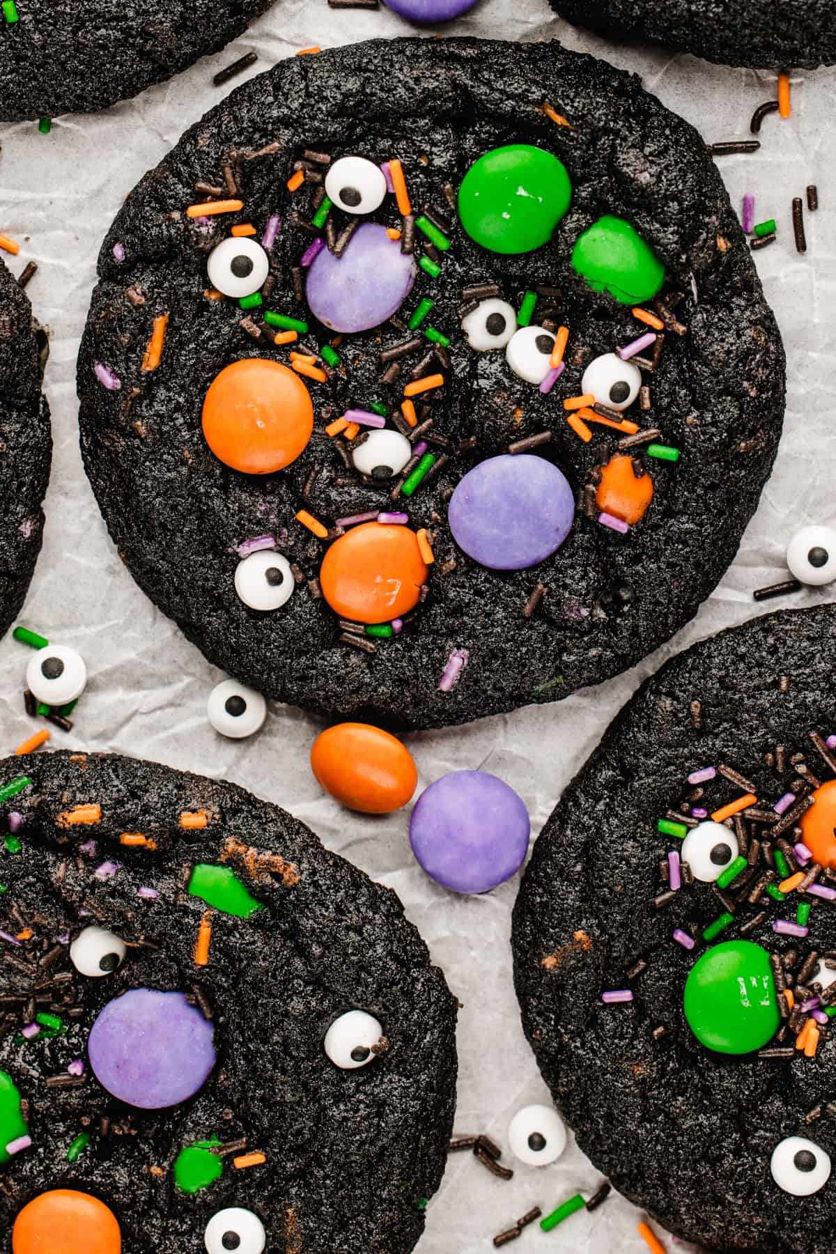 Halloween Cookies Black chocolate cookies decorated with candy eyeballs and orange, purple, and green candy-coated chocolates on parchment paper.