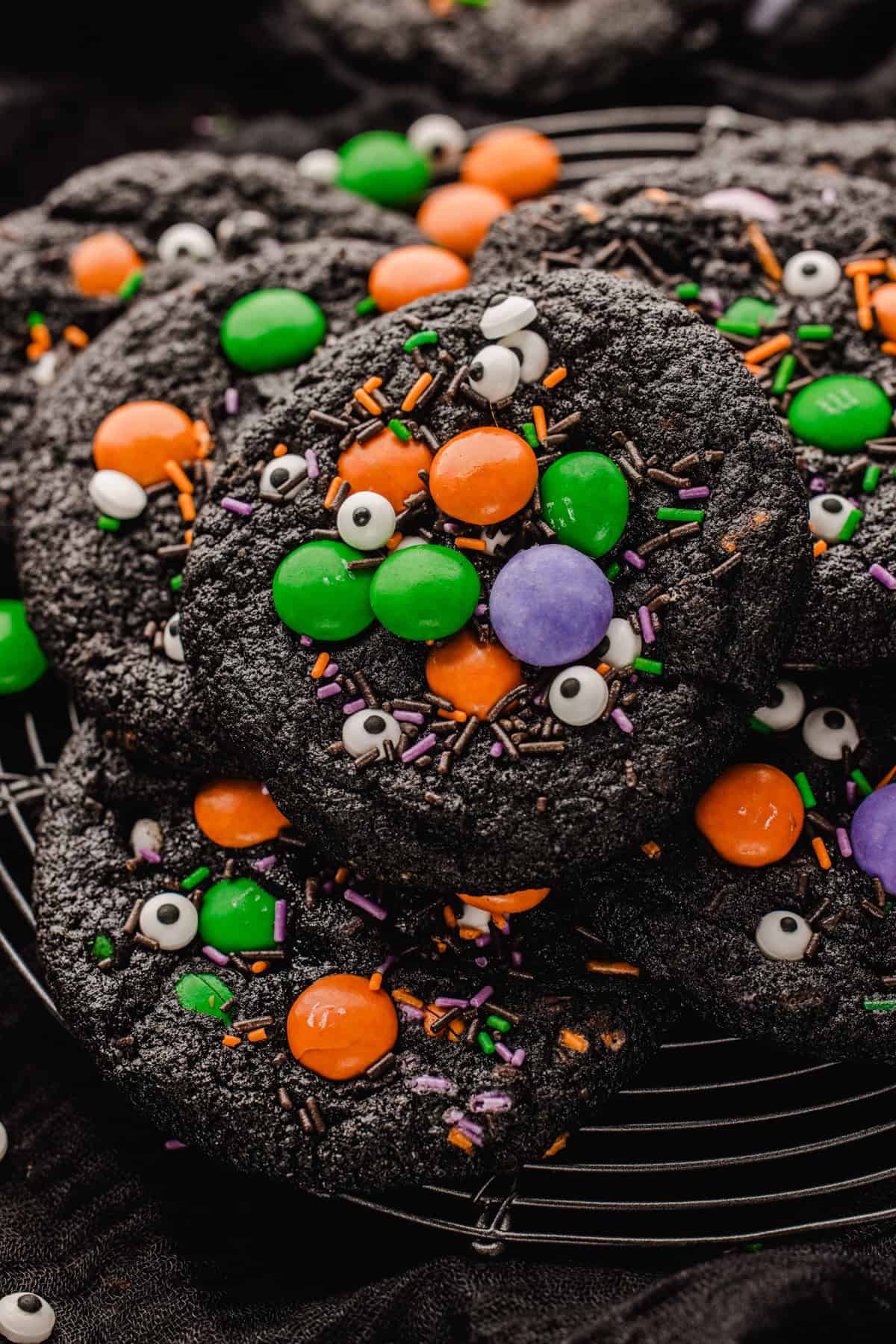Halloween Cookies Black cocoa cookies decorated with colorful candy-coated chocolates, candy eyeballs, and purple and orange sprinkles on a wire cooling rack.