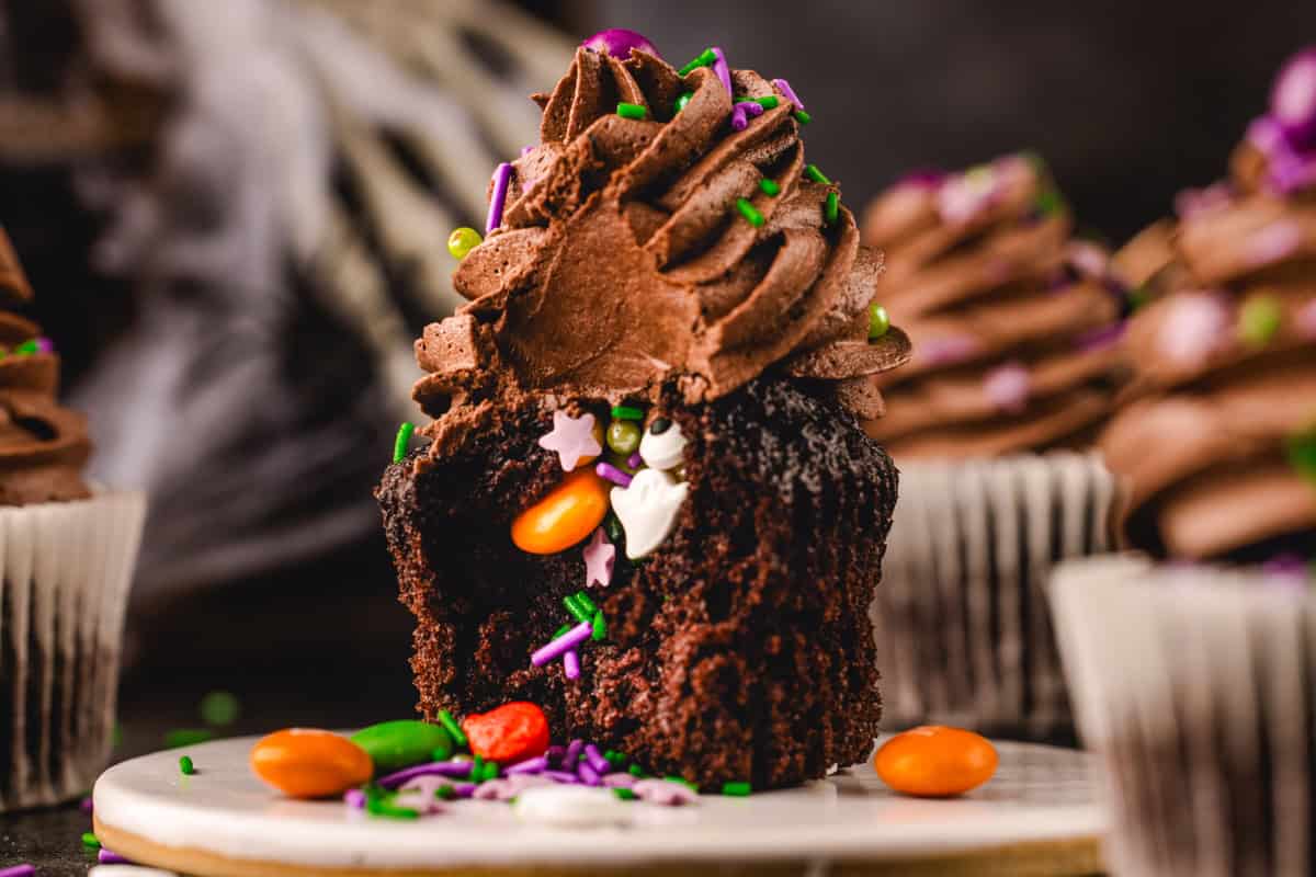 A chocolate cupcake topped with frosting and sprinkles, broken open to reveal a hidden center of  colorful candies.