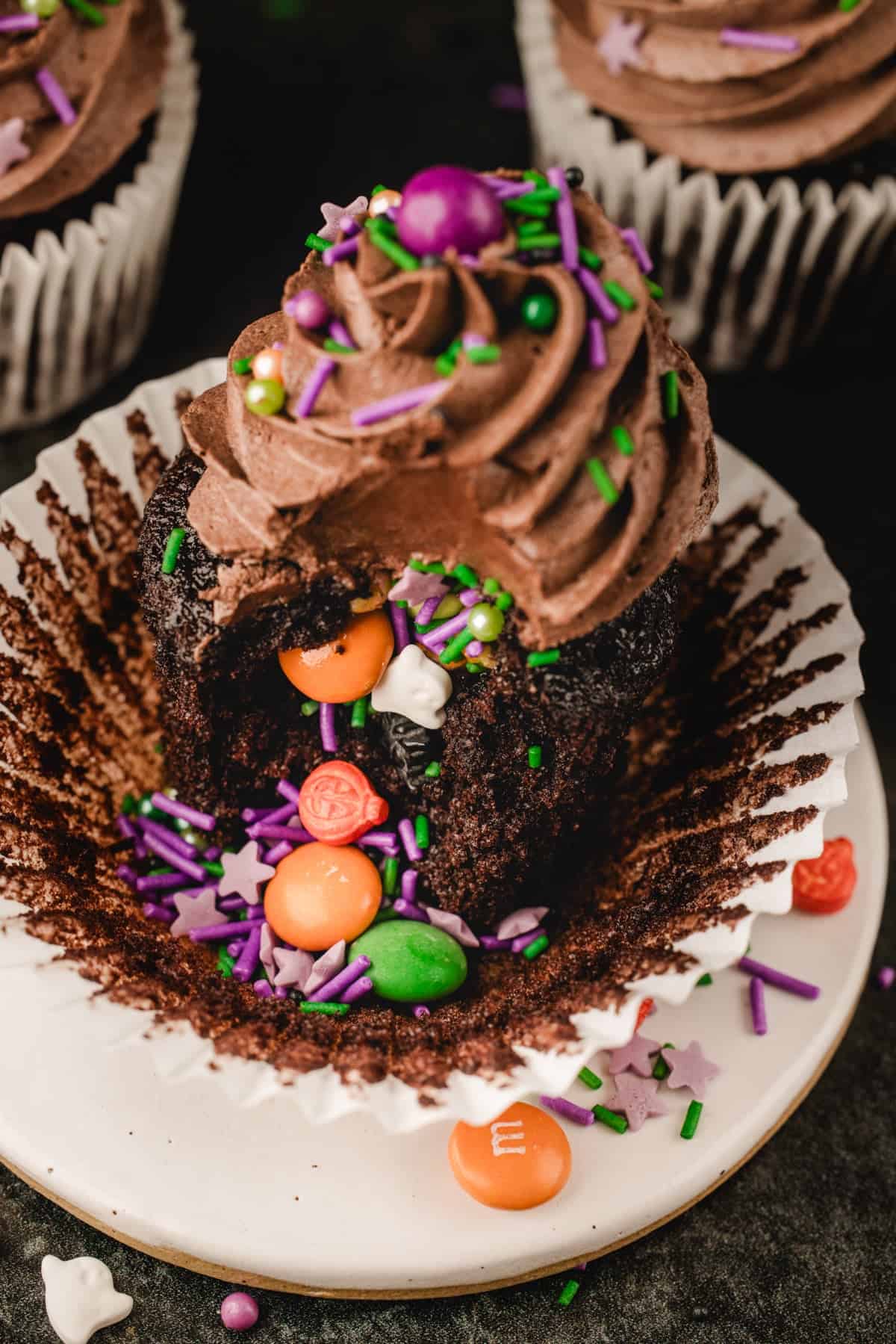 Halloween Cupcakes A chocolate cupcake with chocolate frosting and colorful candy sprinkles, broken open to reveal small candy-coated chocolates inside the center.